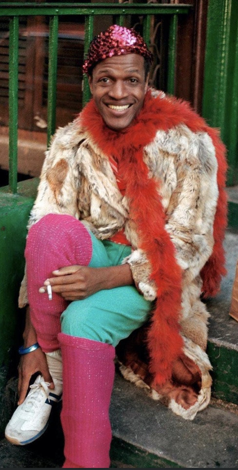 Marsha P. Johnson, shown on this slide in a rare colour photo, is smiling at the photographer, a cigarette in her hand as she sits on a stoop in pink leggings, green pants, a beige patterned jacket, a red scarf, and a sparkly pink hat.