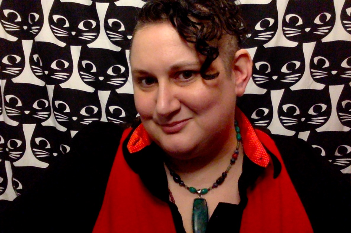 Cat Webb, LET'S low sensory space host, in front of wall of black cats, is wearing a bright red shirt and black sweater.