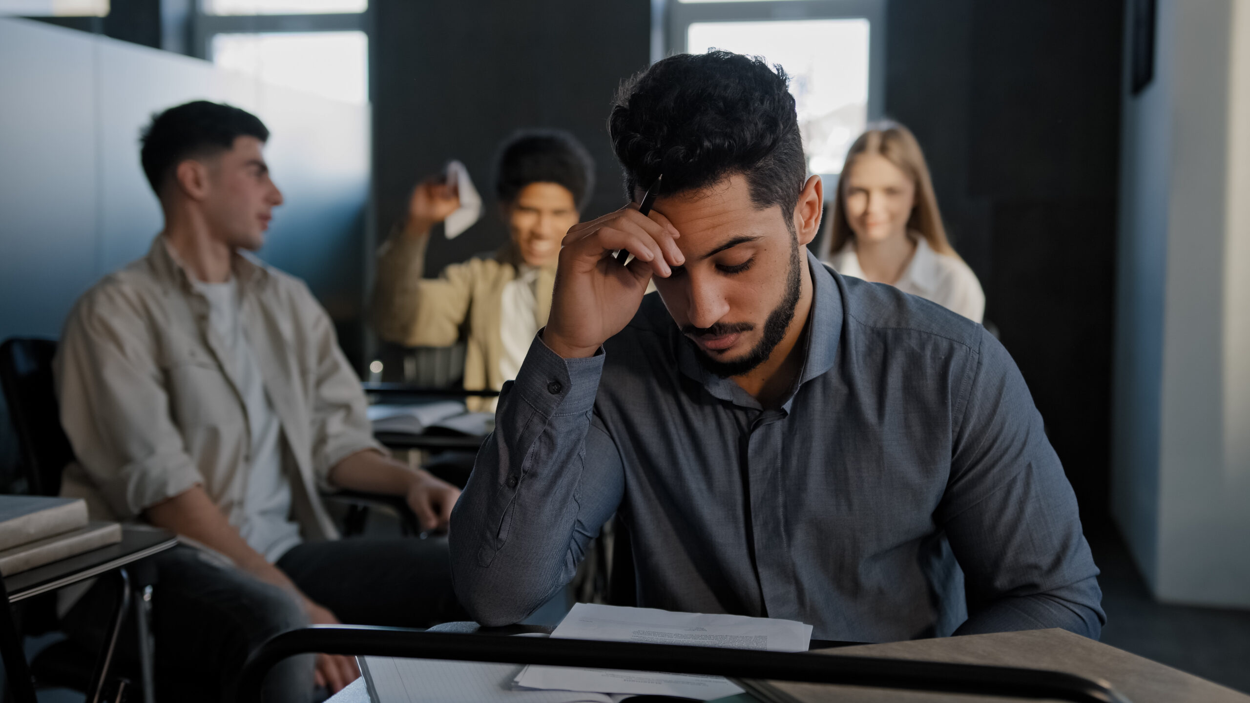 Student, hand on his head, staring down at the desk, alone. A group of happy students are in the background. with a group of students behind them.