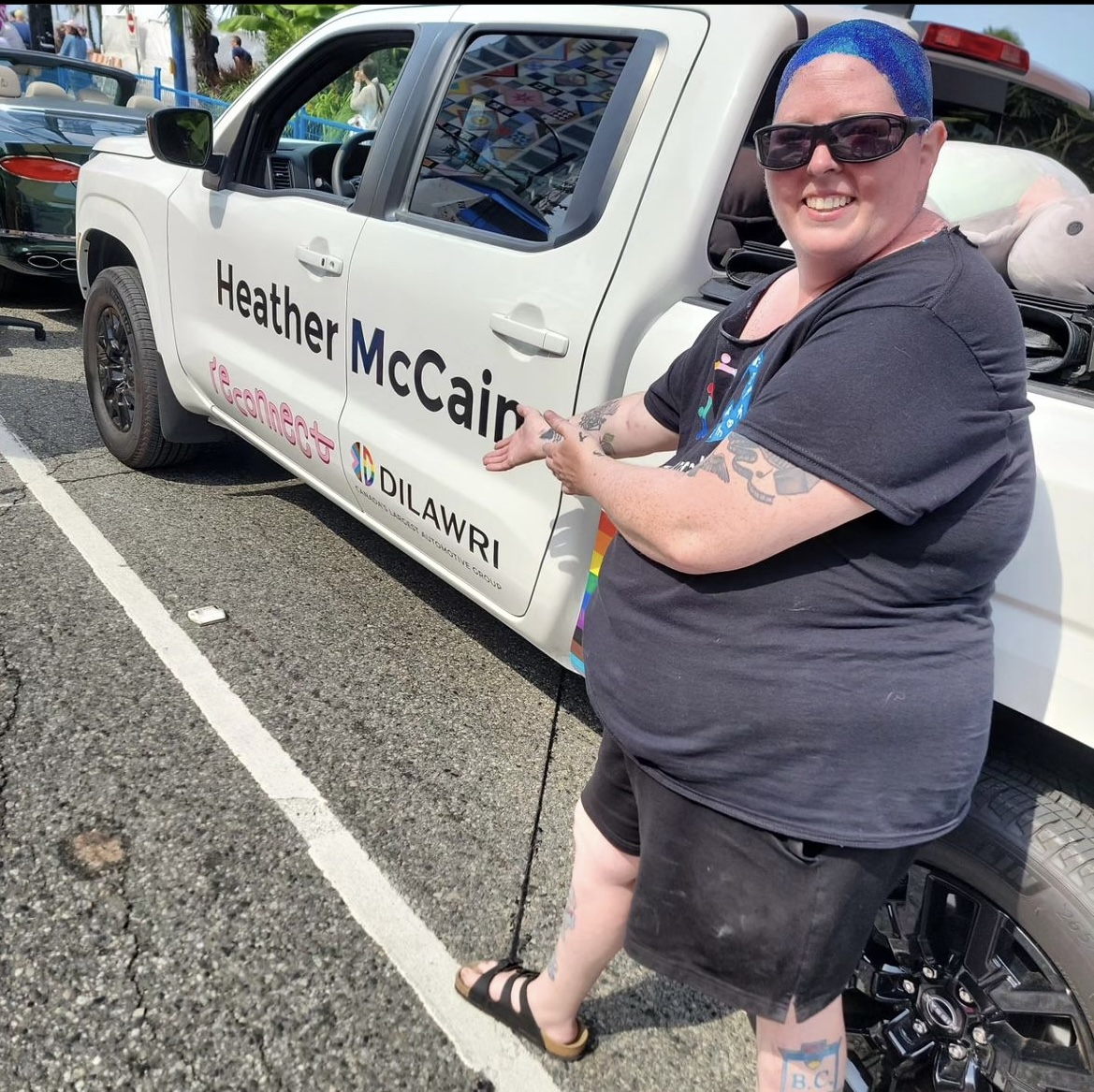 LET'S' Executive Director pointing to a truck with their name. Heather is dressed in all black with their bald head covered completely in blue glitter.