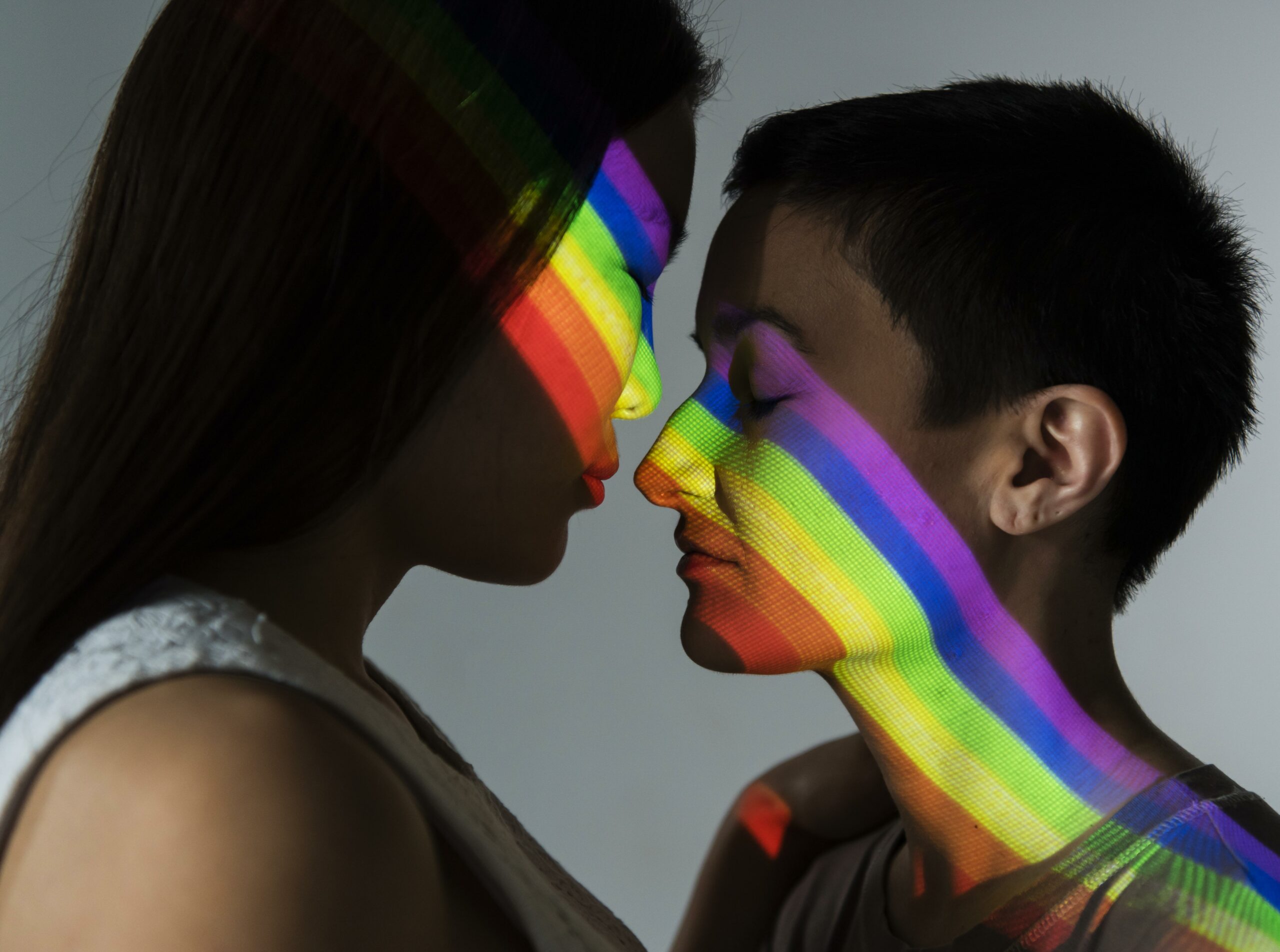 2 people, their noses almost touching, with a rainbow light covering their faces.