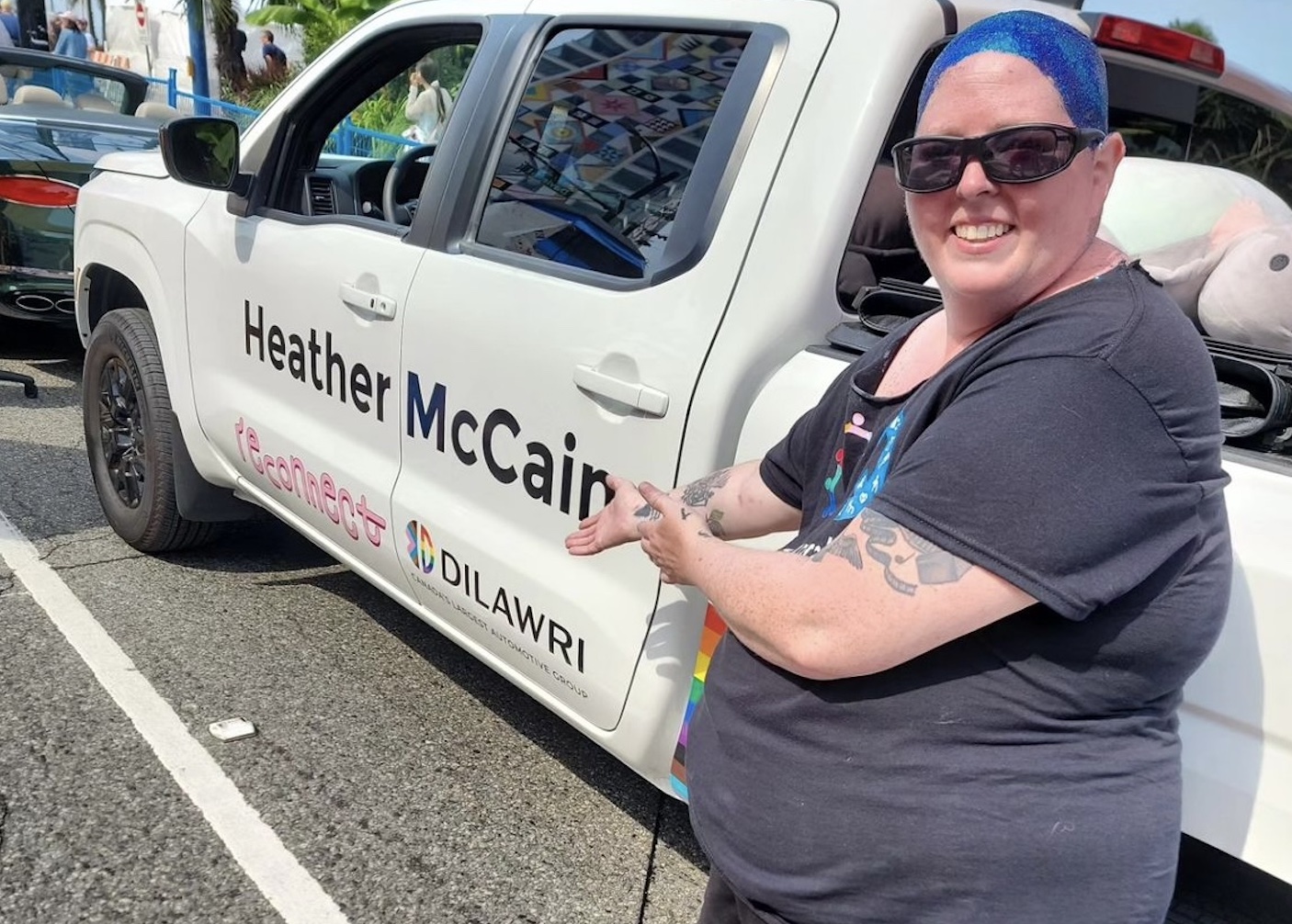 Photo of LET’S’ Executive Director Heather McCain, their head covered in blue glitter, pointing to a truck with their name printed on the side.