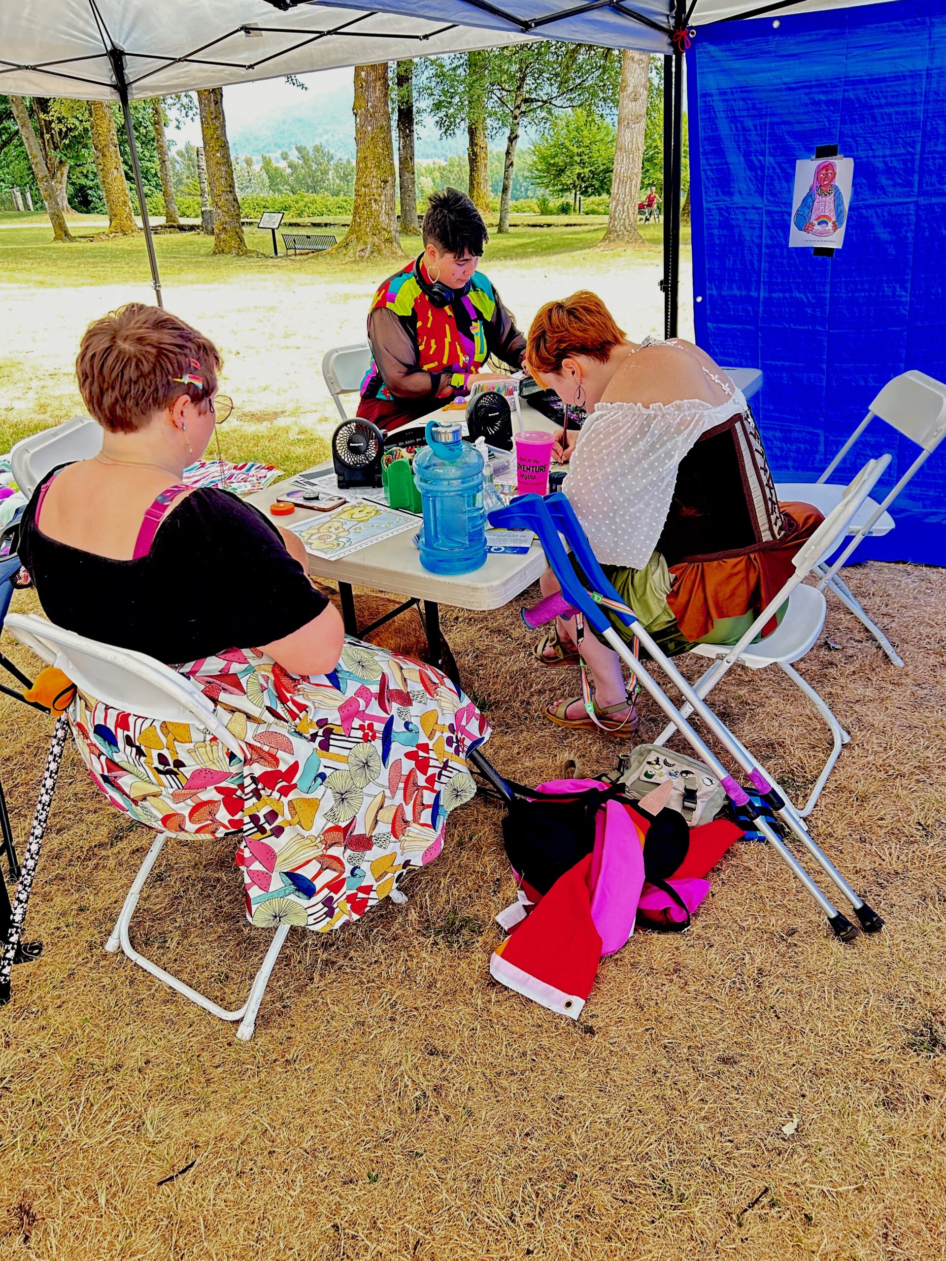 Image: 3 people, 1 with a cane, colouring at a table under a tent in an outdoor space.