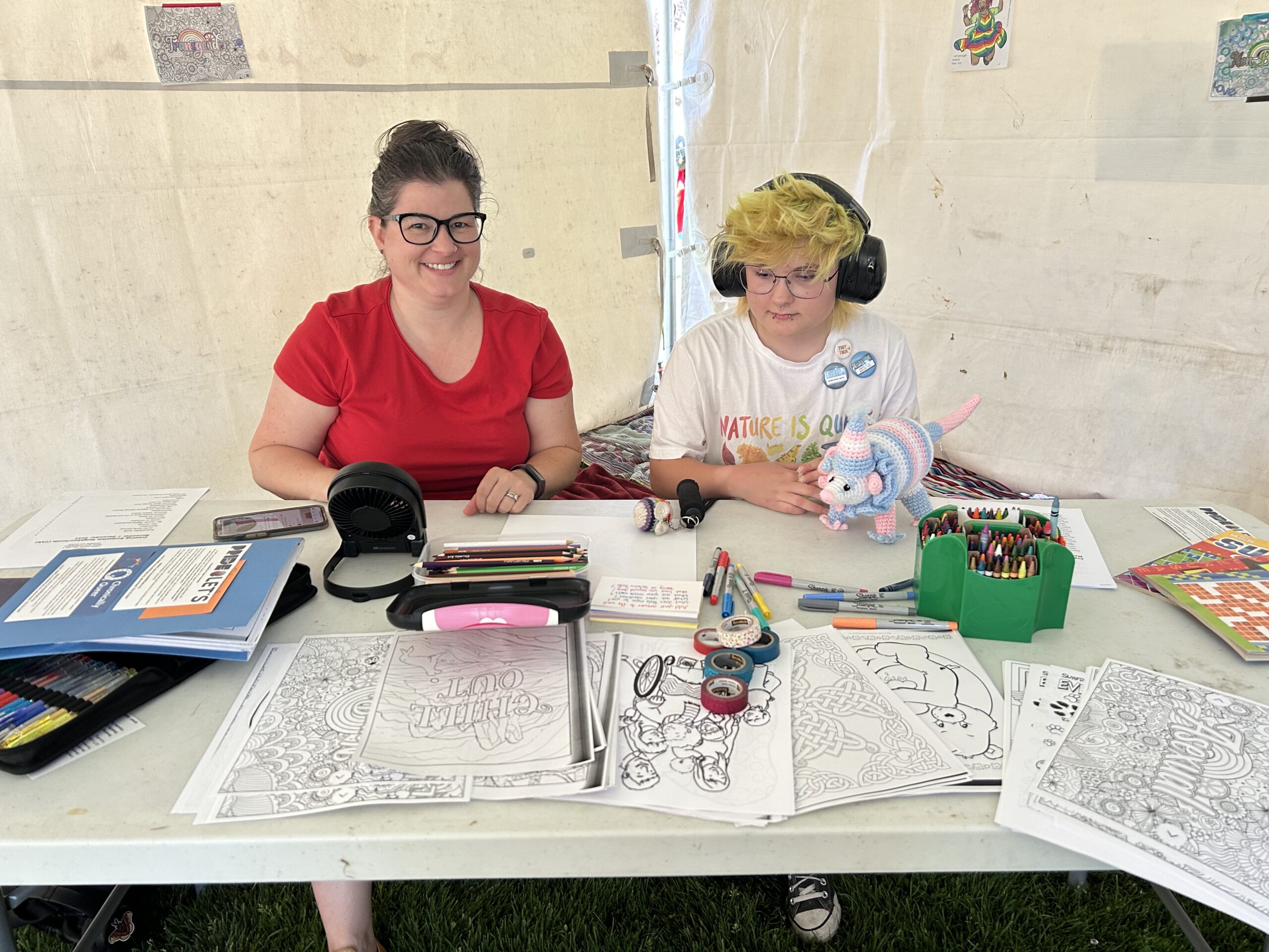 Neurodivergent person, with headphones and a knitted possum in trans colours, and their mum. They are both colouring.