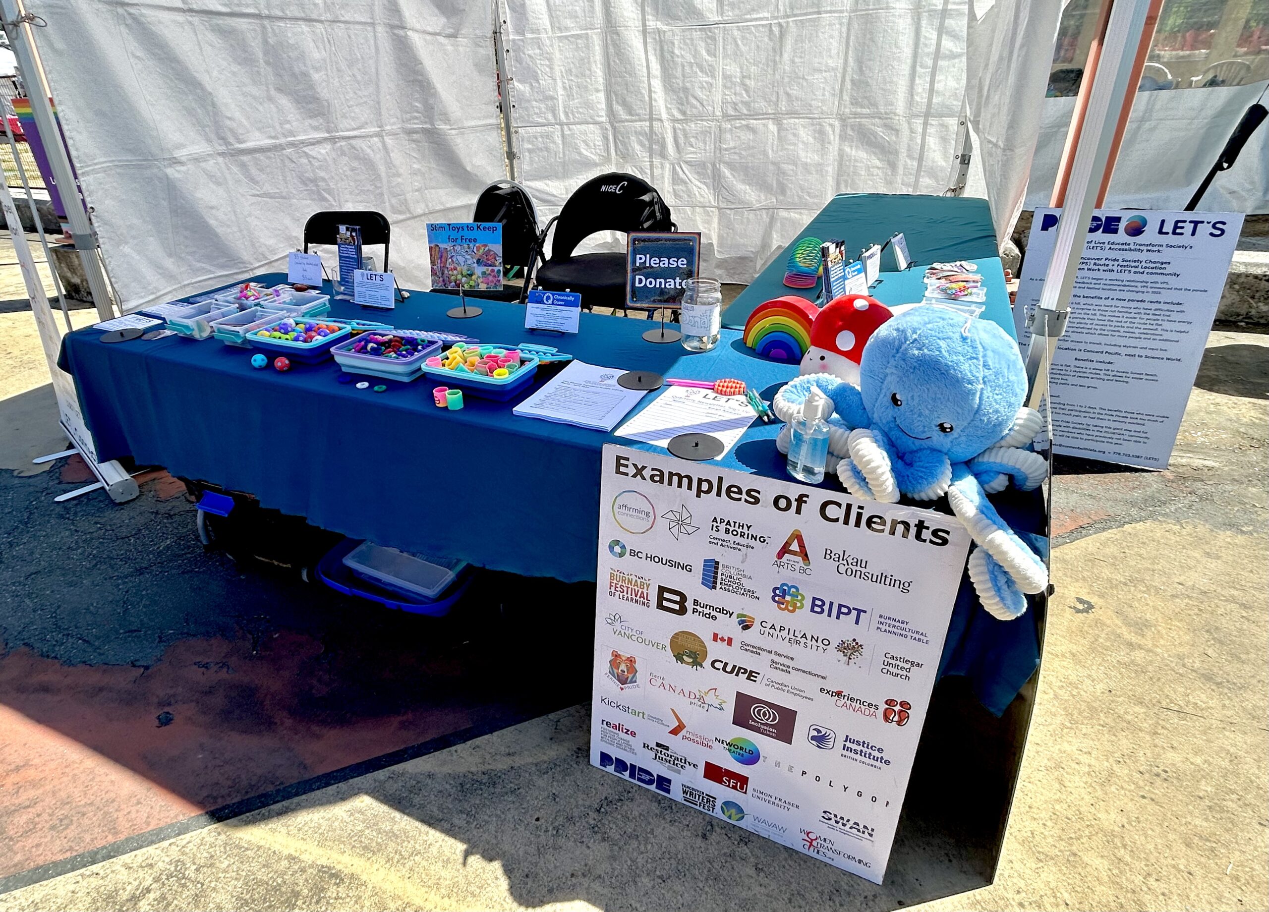 Photo of LET’S’ booth at Vancouver Pride. Our table has stuffies, boxes of stim toys, and signage.