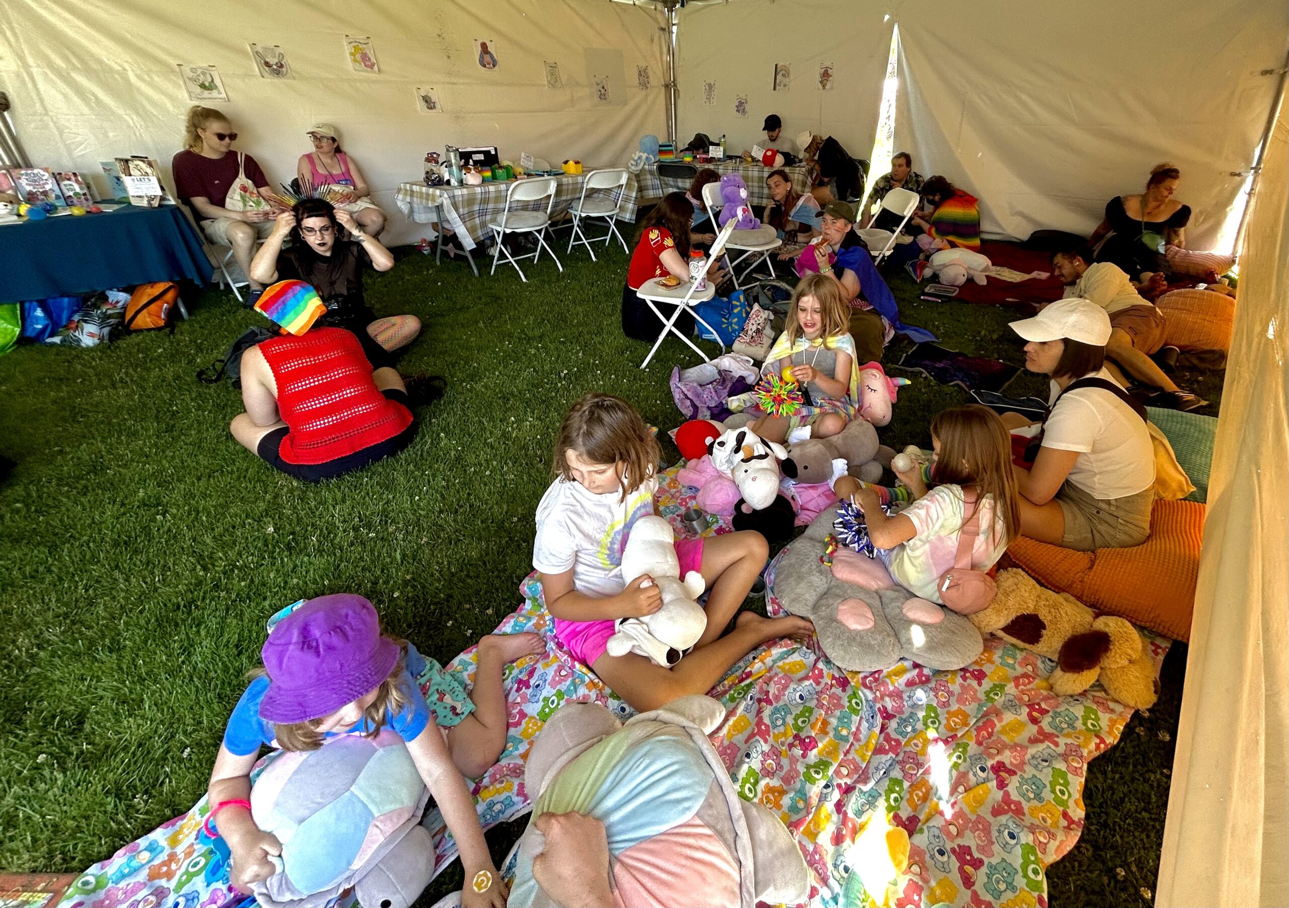 Inside the Victoria Pride tent, people fill the tent, those closest to the viewer are on a Care Bear blanket with stuffed animals.